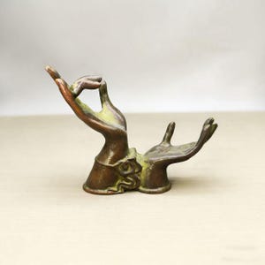 Puede incluir: Escultura de bronce que representa dos manos en un gesto de ofrenda o meditación. Las manos muestran detalles y una pátina verde. La base tiene un diseño ornamentado. Escultura decorativa.