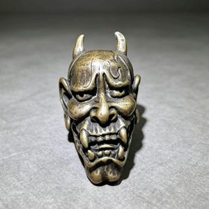May include: A metallic bronze-toned sculpture of a Hannya mask featuring two prominent horns, deep-set eyes, a wide nose, and an open mouth revealing sharp fangs. The surface has a weathered, textured finish with dark patina accents in the crevices.