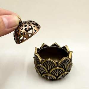 Könnte beinhalten: Ein bronzefarbener Räuchergefäß in Form einer Lotusblume mit einem dekorativen, durchbrochenen Deckel. Der Deckel wird angehoben, um das Innere des Brenners freizulegen. Der Brenner hat ein detailliertes Blütenblattdesign.