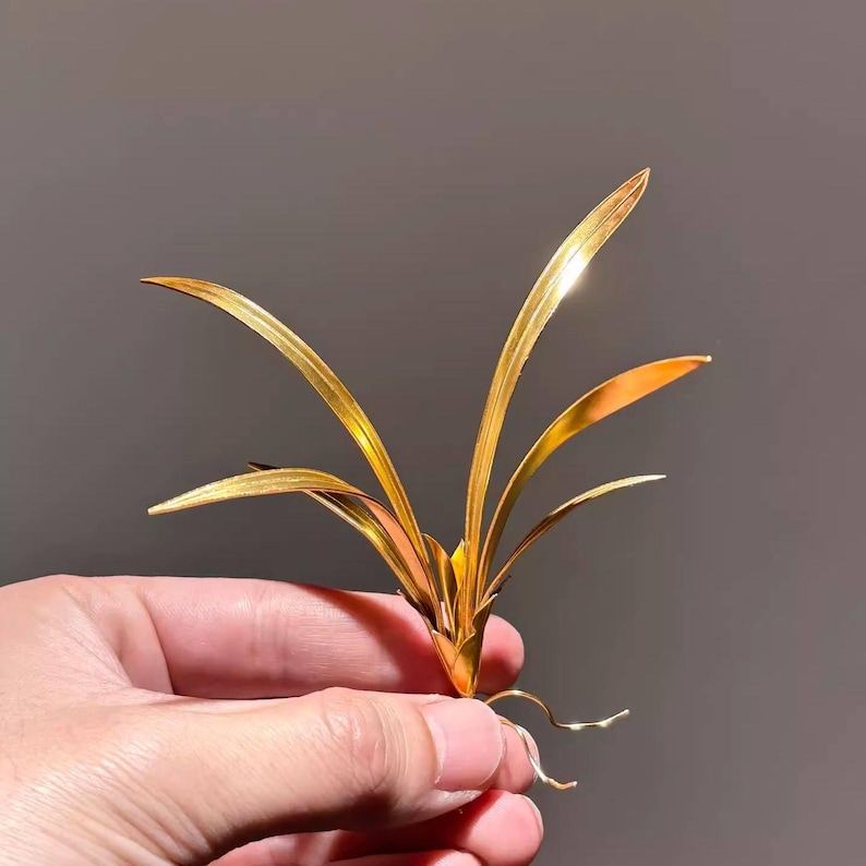 May include: A golden metal plant sculpture with long, slender leaves and delicate roots. The plant is held in a hand against a neutral background. The sculpture has a shiny, reflective surface.