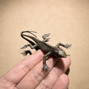 Könnte beinhalten: Detaillierte Bronze-Echsenfigur mit strukturierten Schuppen und ausgestreckten Gliedmaßen. Die Eidechse ist auf einer Hand positioniert und zeigt ihr kompliziertes Design und ihre realistischen Merkmale. Die Figur hat eine dunkle, gealterte Oberfläche.