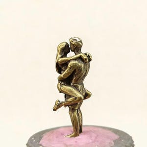 Puede incluir: Una escultura de color bronce de una pareja abraz&aacute;ndose y bes&aacute;ndose. El hombre sostiene a la mujer, que levanta una pierna. La escultura est&aacute; montada sobre una base rosa y gris. Las figuras son detalladas, con rasgos faciales y ropa visibles.