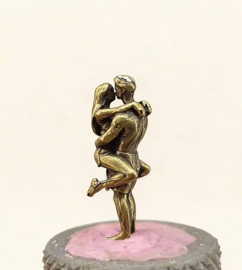 Puede incluir: Una escultura de color bronce de una pareja abraz&aacute;ndose y bes&aacute;ndose. El hombre sostiene a la mujer, que levanta una pierna. La escultura est&aacute; montada sobre una base rosa y gris. Las figuras son detalladas, con rasgos faciales y ropa visibles.