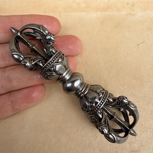 Peut inclure: Un vajra argenté, objet rituel, est tenu dans une main. Le vajra présente des motifs floraux et géométriques ornés, avec une section centrale bulbeuse et des extrémités pointues. L'objet est probablement en métal.