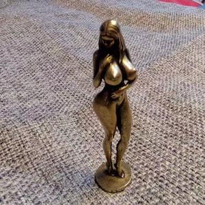 Puede incluir: Una figura de bronce de una mujer en posición de pie. La escultura tiene pelo largo y se agarra los pechos. La figura está sobre una base circular y es de metal.