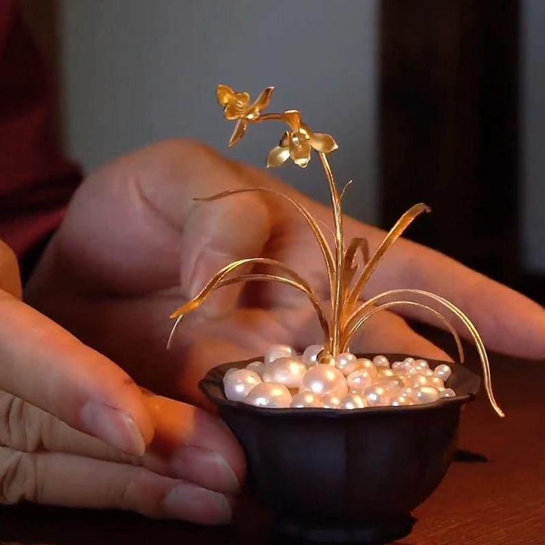 May include: A miniature gold-colored orchid sculpture in a small black pot filled with white pearls. The orchid has delicate petals and long, slender leaves. The sculpture is held in a person's hands.