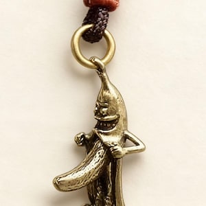 Op de afbeelding: Een messingkleurige hanger in de vorm van een lachend bananenpersonage, met armen en benen, die een banaan vasthoudt. De hanger is bevestigd aan een bruin en zwart koord met een goudkleurige ring.