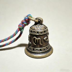 May include: A small, bronze-colored bell with intricate carvings and a colorful braided cord. The bell has a rounded shape with a flared base and a loop at the top for hanging. The cord is made of red, blue, and green threads.