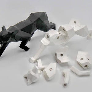 May include: A black geometric panther sculpture and a disassembled white geometric panther sculpture. The black panther is in a stalking pose. The white panther is in pieces, with visible magnets. The sculptures are on a white surface.
