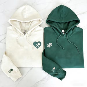 Może przedstawiać: Wyświetlane są dwie bluzy z kapturem, jedna kremowa i jedna zielona. Kremowa bluza ma wzór w kształcie serca z puzzli i literę "B" z sercem na rękawie. Zielona bluza ma element układanki i literę "K" z sercem.