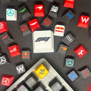 May include: A selection of custom keycaps with diverse logos and designs, such as the Formula 1 logo, Red Bull, and other racing team emblems. The keycaps are in various colours, including red, black, white, and yellow.