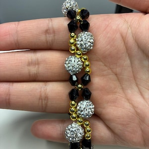 以下が含まれることがあります： 黒と金のビーズが交互に並び、キラキラ光る銀色のラインストーンで覆われたボールが特徴のビーズブレスレット。 ブレスレットは手で持たれており、そのデザインと色のコントラストが際立っています。