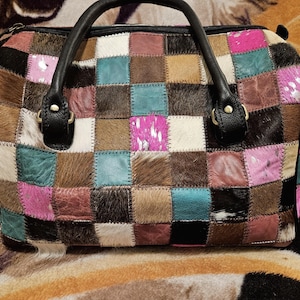 May include: A patchwork handbag with a variety of colors and textures. The bag features a checkerboard pattern of squares in shades of brown, pink, teal, and white. It has black leather handles and trim. The bag is sitting on a patterned surface.