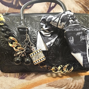 May include: A black barrel-shaped handbag with an embossed floral pattern, featuring a black and white patterned scarf tied to the handle, a gold and black chain strap, and decorative charms including two black cherries and a small perfume bottle.