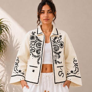 May include: An off-white jacket featuring black embroidered botanical and celestial motifs. The jacket has a collared neckline, long sleeves with flared cuffs, and two front pockets. The embroidery includes floral patterns, stars, and moons.