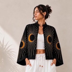 May include: A black open-front jacket featuring gold embroidered sun and moon motifs on the shoulders, paired with a white cropped tank top and white wide-leg trousers with a drawstring waist and wooden bead accents at the ends of the ties.