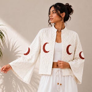 Può includere: Una giacca color crema aperta sul davanti con maniche larghe, caratterizzata da motivi ricamati a mezzaluna rosso scuro e linee radianti. La giacca è indossata sopra un top corto bianco e pantaloni bianchi a gamba larga con coulisse.