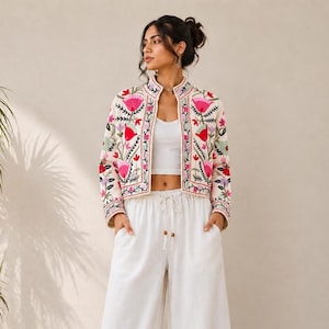 Peut inclure: Une veste courte couleur crème ornée de broderies florales vives dans des tons rose, rouge et vert, associée à un haut court blanc uni et un pantalon blanc à jambes larges et taille haute avec un cordon de serrage et des perles en bois.