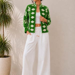 May include: A vibrant green jacket featuring a white floral and geometric pattern, worn over a white cropped tank top and loose-fitting white wide-leg trousers with a drawstring waist and wooden bead accents at the ends of the cord.