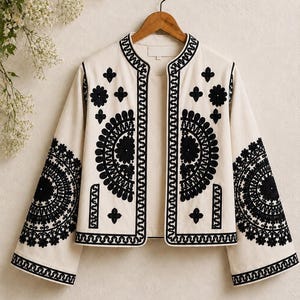 May include: An off-white jacket featuring black embroidery in circular and floral patterns. The garment has a mandarin collar, long wide sleeves, and a straight hemline, with black embroidered trim along the front opening and sleeve cuffs.