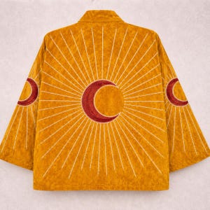 May include: A mustard yellow jacket featuring a large, embroidered dark red crescent moon at the centre of the back, surrounded by radiating white embroidered sunburst lines that extend across the back and onto both sleeves.