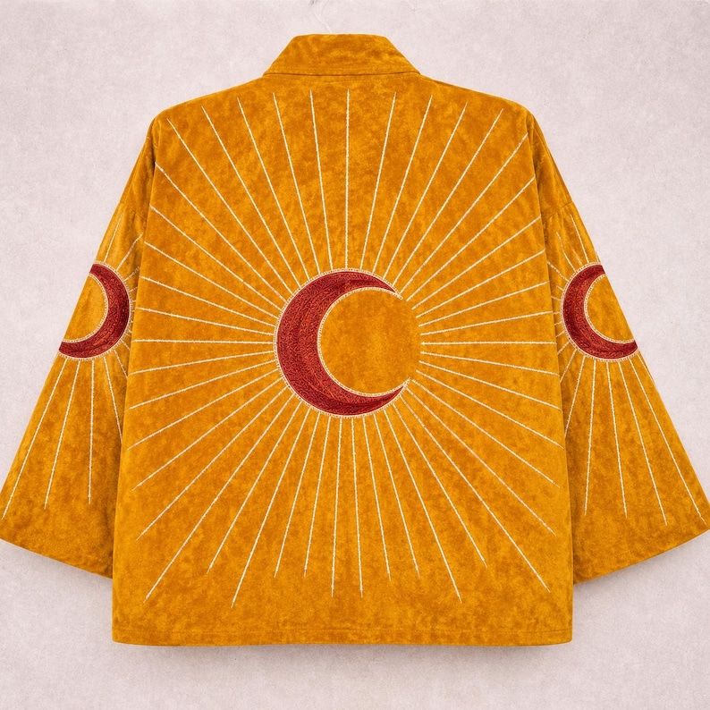 May include: A mustard yellow jacket featuring a large, embroidered dark red crescent moon at the centre of the back, surrounded by radiating white embroidered sunburst lines that extend across the back and onto both sleeves.