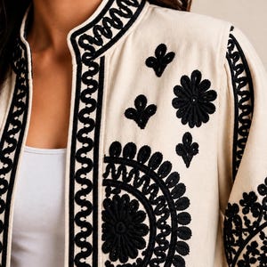 May include: A cream-colored jacket featuring intricate black embroidery along the front opening, collar, and sleeves. The design includes bold geometric wave patterns and detailed floral motifs, creating a high-contrast visual effect.