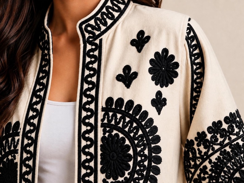 May include: A cream-colored jacket featuring intricate black embroidery along the front opening, collar, and sleeves. The design includes bold geometric wave patterns and detailed floral motifs, creating a high-contrast visual effect.
