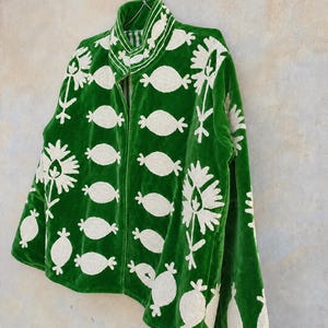 May include: A vibrant green velvet jacket featuring a high collar and a repeating pattern of cream-colored embroidered floral and oval motifs. The garment has a front zipper closure and long sleeves, showcasing detailed needlework throughout the fabric.