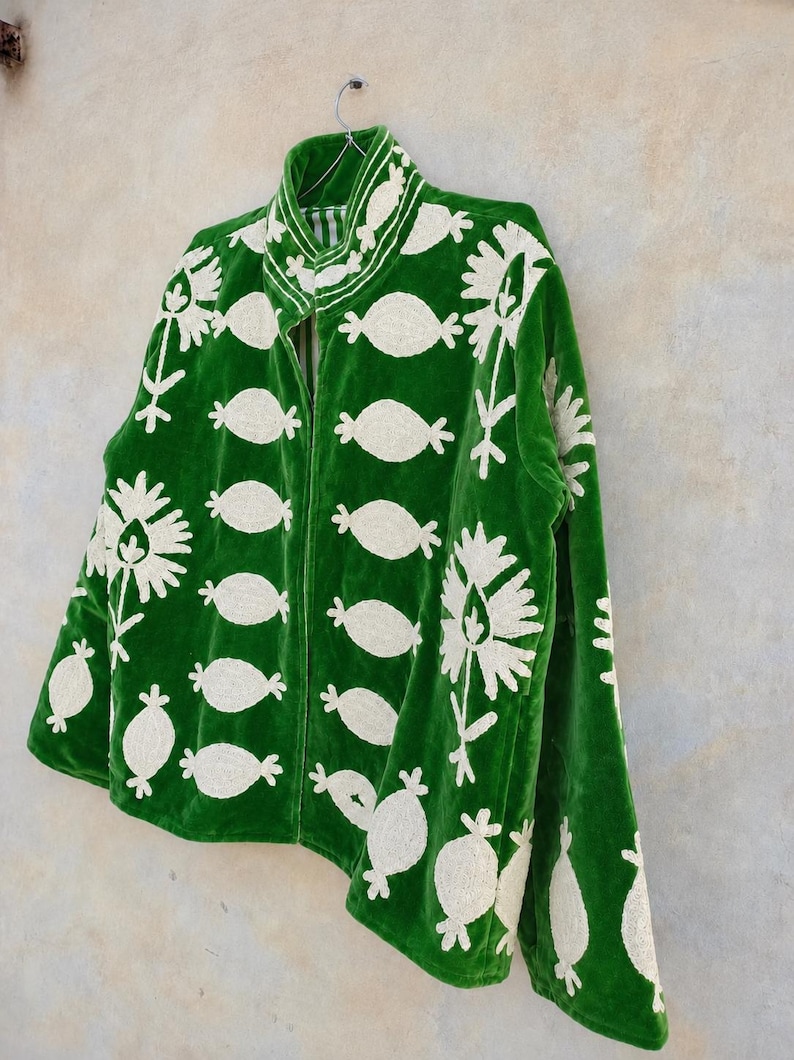 May include: A vibrant green velvet jacket featuring a high collar and a repeating pattern of cream-colored embroidered floral and oval motifs. The garment has a front zipper closure and long sleeves, showcasing detailed needlework throughout the fabric.