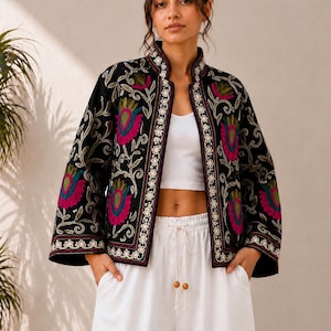 Könnte beinhalten: Eine schwarze, vorne offene Jacke mit Stehkragen und weiten Ärmeln. Das Kleidungsstück ist mit aufwendigen Blumenstickereien in leuchtendem Magenta, Grün und Weiß verziert, wobei ein sich wiederholendes weißes Blumenmuster die Kanten säumt.