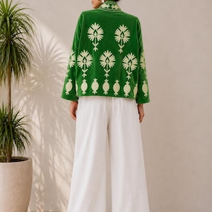 May include: A vibrant green jacket featuring a light cream-colored floral embroidery pattern on the back and sleeves, paired with loose-fitting white wide-leg trousers. The jacket has a high collar and long sleeves, creating a coordinated outfit.