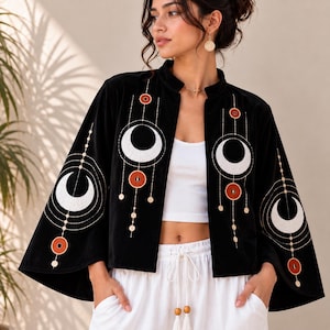 Könnte beinhalten: Eine schwarze Jacke mit offener Vorderseite und weiten Ärmeln, verziert mit weißen Halbmondstickereien und kreisförmigen orangefarbenen Akzenten. Das Design umfasst feine weiße Linien, die geometrische Muster um die Monde bilden, getragen über einem weißen bauchfreien Oberteil.