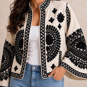 May include: A cream-colored jacket featuring a mandarin collar and wide sleeves, adorned with intricate black embroidery in circular and floral patterns along the front panels and sleeves, worn over a white top and blue denim jeans.