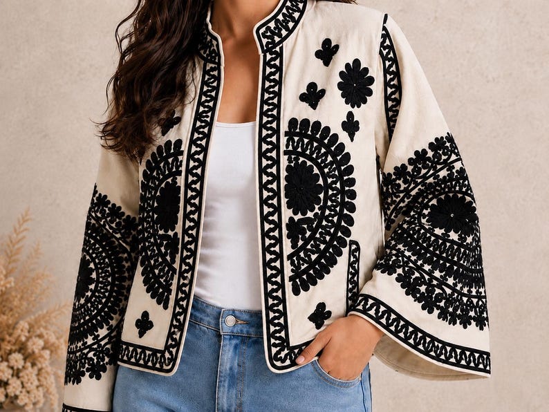 May include: A cream-colored jacket featuring a mandarin collar and wide sleeves, adorned with intricate black embroidery in circular and floral patterns along the front panels and sleeves, worn over a white top and blue denim jeans.