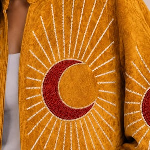May include: A mustard yellow jacket featuring a large embroidered red crescent moon at the centre, surrounded by radiating white stitched sunburst rays. The fabric has a textured, crushed appearance, and the design is repeated on the sleeve.