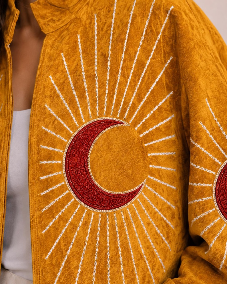 May include: A mustard yellow jacket featuring a large embroidered red crescent moon at the centre, surrounded by radiating white stitched sunburst rays. The fabric has a textured, crushed appearance, and the design is repeated on the sleeve.