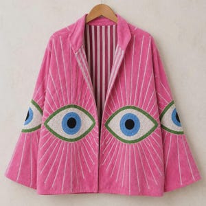 Puede incluir: Una chaqueta rosa brillante abierta por delante con grandes motivos de ojos bordados en el frente y las mangas. Los ojos tienen iris azules, pupilas negras y esclerótica blanca con bordes verdes. El forro interior presenta rayas verticales.