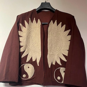 Könnte beinhalten: Eine dunkelbraune, offene Jacke mit großen, cremefarbenen, gestickten Sonnenstrahlen-Motiven auf der Brust und zwei Vordertaschen, die jeweils mit einem cremefarbenen, gestickten Yin-Yang-Symbol verziert sind. Die Jacke hat lange Ärmel und einen kragenlosen Ausschnitt.