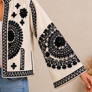 May include: An off-white jacket featuring intricate black embroidery. The design includes large circular floral patterns on the sleeves and front panels, a mandarin collar, and wide sleeves. The embroidery consists of dense, dark stitching.