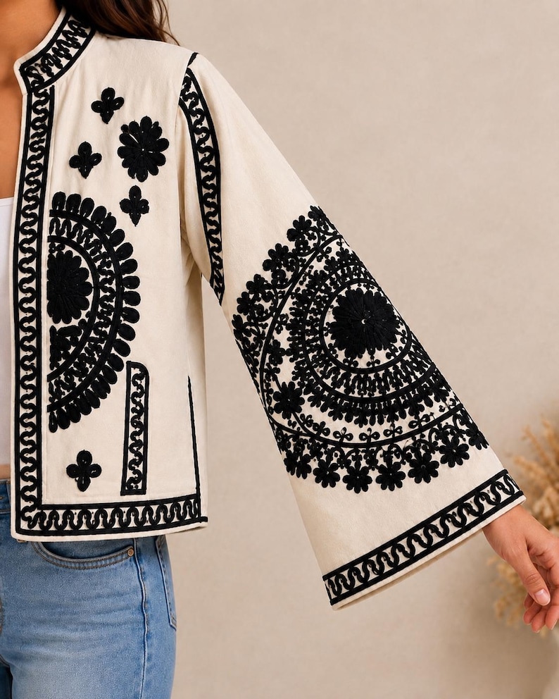 May include: An off-white jacket featuring intricate black embroidery. The design includes large circular floral patterns on the sleeves and front panels, a mandarin collar, and wide sleeves. The embroidery consists of dense, dark stitching.