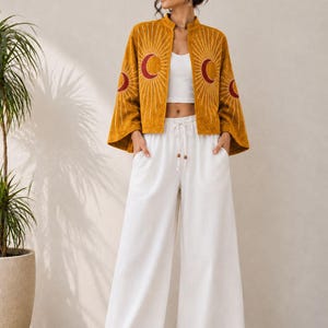 May include: A mustard yellow open-front jacket featuring embroidered red crescent moons and white sunburst rays on the sleeves and chest, paired with a white cropped tank top and white wide-leg trousers with a drawstring waist and wooden beads.