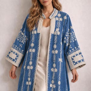 May include: A long, open-front blue coat with intricate white floral embroidery. The coat features a cream-colored trim along the edges and sleeves. The sleeves are wide and the coat is knee-length, creating a bohemian aesthetic.