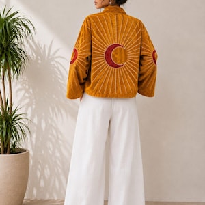 May include: A mustard yellow velvet jacket featuring a large embroidered red crescent moon on the back with white radiating sunburst lines, and smaller matching moon motifs on each sleeve, paired with loose-fitting white wide-leg trousers.