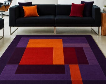May include: A rectangular area rug with a geometric design in shades of purple, orange, and red. The rug features a series of layered rectangles and squares, creating a modern, abstract pattern. The rug is placed in a living room setting with a black sofa and colorful throw pillows.