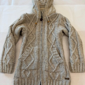 May include: A beige cable-knit hooded cardigan featuring a full-length front zipper, long sleeves with ribbed cuffs, and two front pockets. The knit pattern includes vertical diamond shapes, and the hood has two small wooden beads on drawstrings.