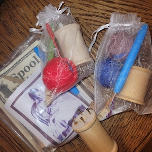 May include: A collection of craft supplies, including wooden spools, yarn, and needles, presented in small mesh bags. A red yarn ball and a printed image are also visible. The items are arranged on a wooden surface.