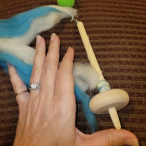 May include: A wooden drop spindle with a light blue and white fiber bundle. The spindle has a mushroom-shaped whorl and a wooden shaft. Two green and one light green pom-poms are attached to the top.