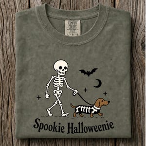 May include: An olive green t-shirt featuring a graphic of a white skeleton walking a brown dachshund wearing a skeleton costume. The design includes a black bat, a crescent moon, and stars. The text Spookie Halloweenie is printed below the graphic.