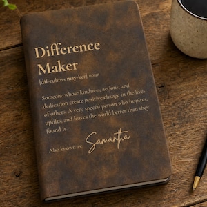 Pode incluir: Um caderno castanho escuro com acabamento texturizado apresentando o texto dourado Difference Maker, uma pronúncia fonética, uma definição do termo, a frase Also known as e o nome Samantha escrito numa fonte de escrita cursiva.